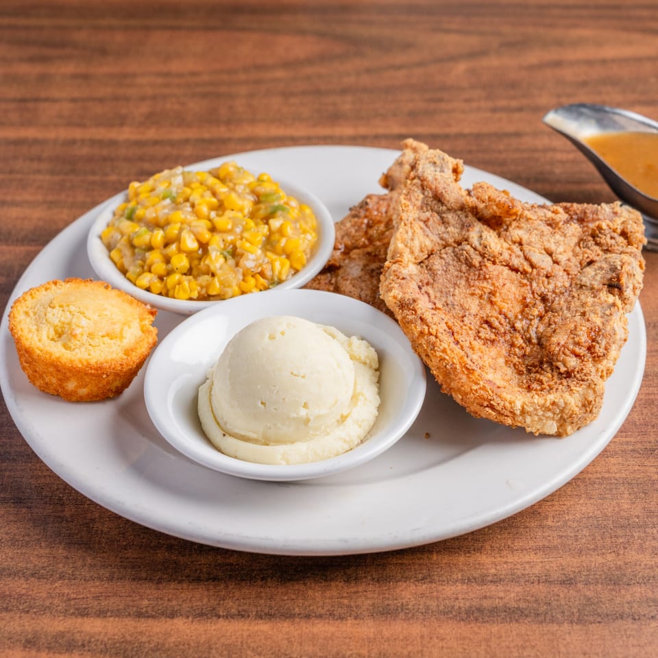 Fried pork chop, creamed corn, cornbread, and mashed potatoes with gravy on a white plate.
